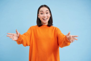 Dost canlısı Asyalı kadın elini tutmak için uzanır, mutlu bir gülümsemeyle karşılar. Kadın model kollarını uzatıp sarılıyor, mavi arka planda duruyor..