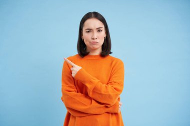 Şüpheli genç Asyalı kadın, parmağıyla reklamı işaret ediyor ve şüpheci bir ifadeyle, şüpheli görünüyor, mavi arka planda duruyor..