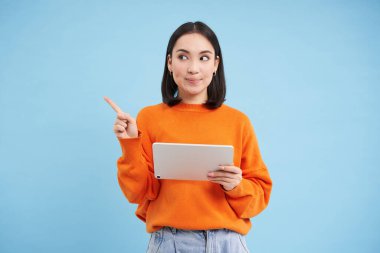 Soldaki boş fotokopi alanını gösteren Asyalı genç bir kadın resmi, dijital tablet tutuyor ve düşünüyor, online alışveriş kavramı, mavi arka plan.