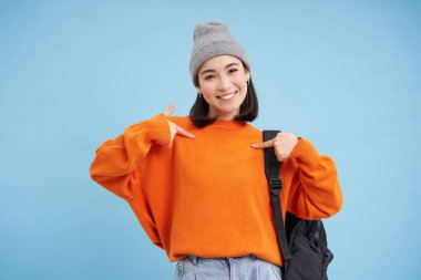 Tarz sahibi Koreli kadın kendini işaret ediyor, sıcak bir şapkayla duruyor ve sırt çantasıyla mavi arka planda duruyor. Boşluğu kopyala