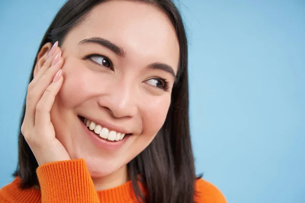 Beauty and skincare. Happy asian woman touches her skin, clear healthy face without blemishes, smiling, standing over blue background.