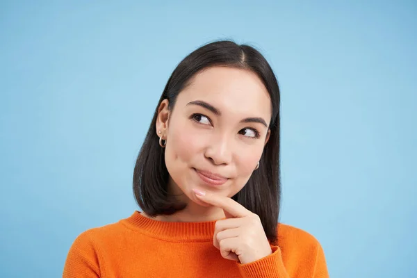 stock image Close up of thinking asian girl, looks pleased and touches chin thoughtful, stands over blue background.