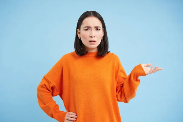 So what. Frustrated and confused asian woman shrugs, raises one hand and looks puzzled, dont understand, stands clueless against blue background.