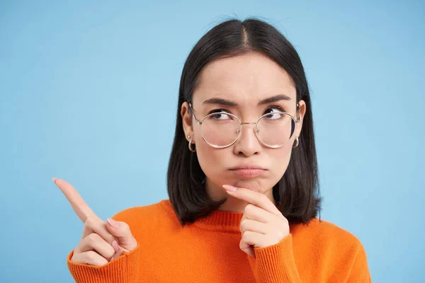 Image of thoughtful asian woman frowns, wears glasses, points left and thinks, makes serious decision, blue background.