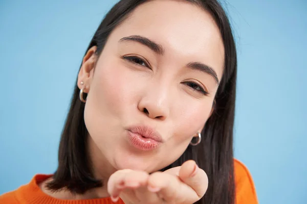 Close up of beautiful korean woman, blowing air kiss at camera, standing over blue background.