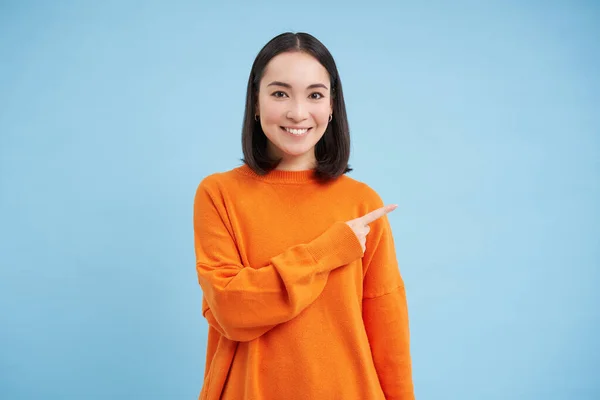 stock image Happy smiling asian woman, pointing finger to the right, shows advertisement, blank copy space on blue background.