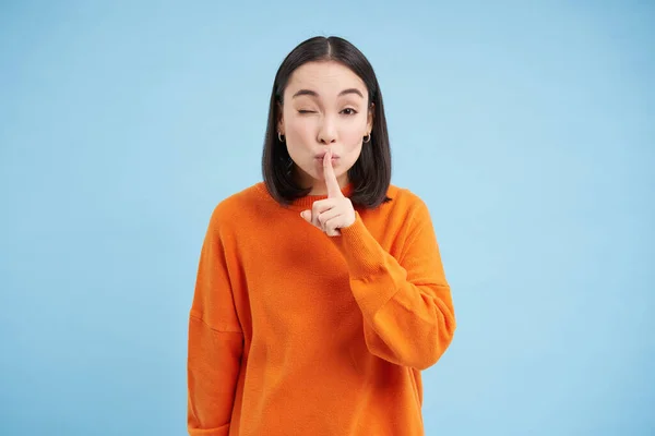 Hush its a secret. Young asian woman shushing, asks to be quiet, holds finger on lips, gossips, stands in orange sweatshirt over blue background.