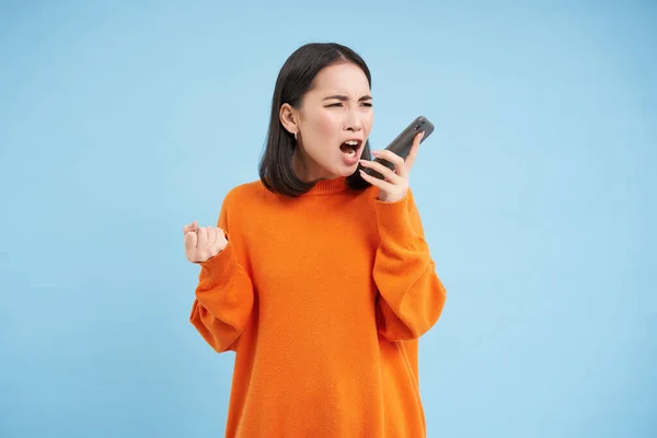 怒气冲冲的亚洲女人对着手机大喊大叫 在智能手机里怒气冲冲地大叫 站在蓝色的背景上 — 图库照片