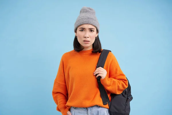 stock image Young asian woman in hat, holds backpack, sulks and looks confused, perplexed emotion, stands over blue background.