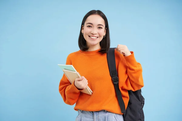 Stock image Education and students. Smiling young asian woman with backpack and notebooks, posing against blue background.