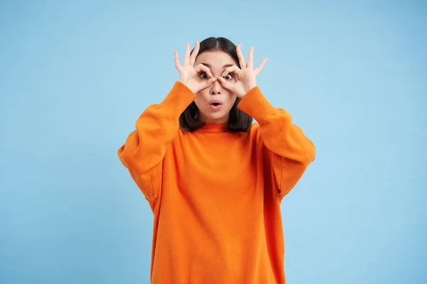 Wow, look at that. Portrait of amazed young asian woman, looks through hand glasses with impressed face expression, stands over blue background.