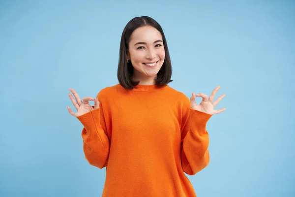 Everything ok. Smiling korean female model, shows okay, alright gesture, approves smth, gives positive feedback, says yes, stands over blue background.