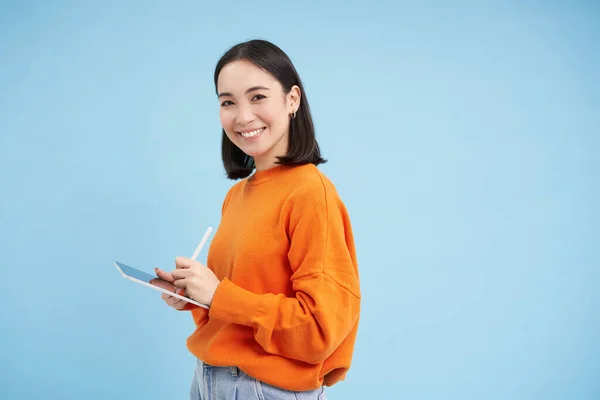 stock image Portrait of young Chinese woman, teacher or student with digital tablet and pencil, writing, taking notes, doing her homework, standing over blue background.