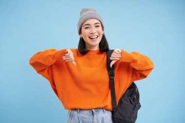 Mutlu gülen kadın, mavi arka planda sırt çantasıyla poz veren kaygısız bir yüz ifadesiyle baş parmağını gösteriyor..