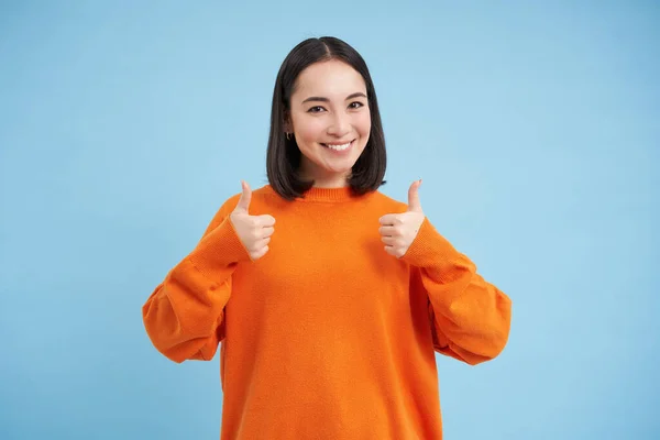 Good job. Smiling korean woman shows thumbs up in approval, like smth, gives good feedback, nods in positive reply, recommends something, blue background.