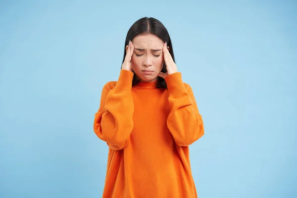 stock image Frustrated asian woman holds hands on head, has headache, looks miserable, cant handle pressure, feels distressed, stands over blue background. Mental health concept.