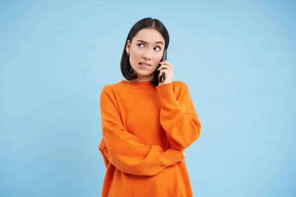Girl with puzzled face answers phone call, looks confused while talks on cellphone, stands over blue background.