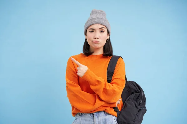 stock image Skeptical asian woman pointing left at strange banner, looks doubtful, stands over studio background.