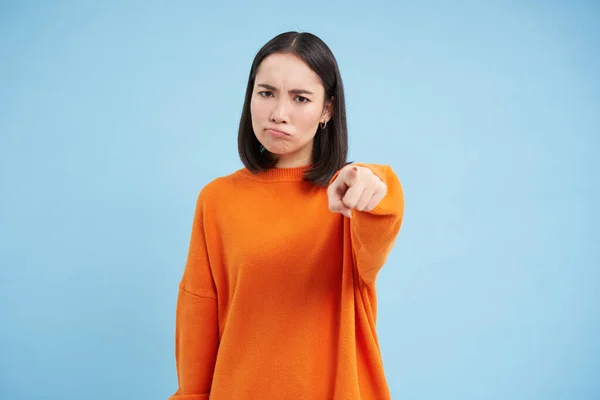 Angry frowning asian woman pointing finger, accusing, blaming someone, being disappointed, standing over blue background.