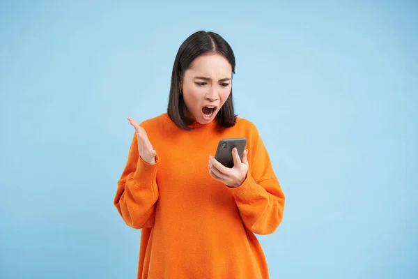 怒气冲冲的韩国女孩一边在手机上咒骂着 一边怒气冲冲地看着智能手机上的新闻 站在蓝色的背景上 — 图库照片