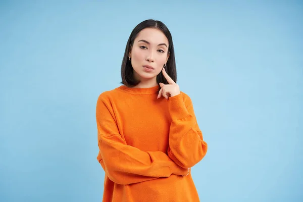 stock image Thoughtful asian woman, looking with interest at camera, thinking smth, making decision, standing over blue background. Copy space