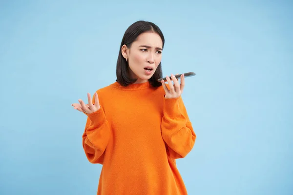 Portrait of upset young woman records voice mail or message, complains with disappointed, sad face, standing over blue background and holding mobile phone.