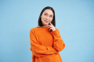 Gülümseyen Asyalı kadın, düşünceli, mutlu bir yüz ifadesiyle yukarı bakıyor, ilginç bir fikri var, bir şeyler planlıyor, mavi arka planda duruyor..