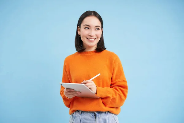 stock image Portrait of young Chinese woman, teacher or student with digital tablet and pencil, writing, taking notes, doing her homework, standing over blue background.