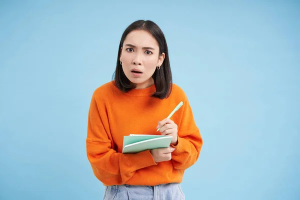 Portrait of confused asian girl, taking notes and looking puzzled while writing, standing over blue background.