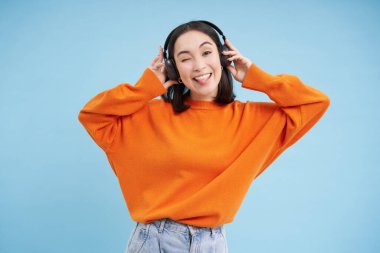 Kulaklıklı mutlu Çinli kadın, müzik dinliyor, müzik listesindeki en sevdiği şarkıyı seviyor, mavi arka planda duruyor. Boşluğu kopyala