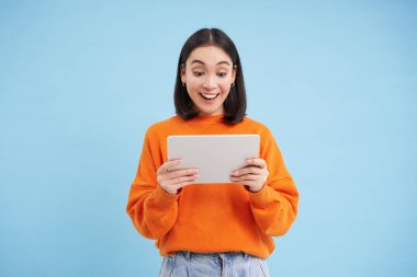 Gülümseyen Asyalı güzel bir kadın dijital tablette okuyor, cihaz videoları izliyor ya da video oyunları oynuyor, mavi arka planda duruyor..