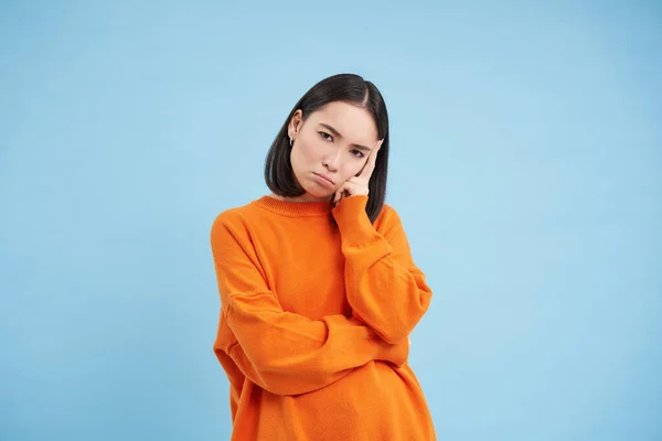 Sad sulking girl leans on hand, looks disappointed and frustrated, angry at someone, stands in orange sweatshirt over blue background.