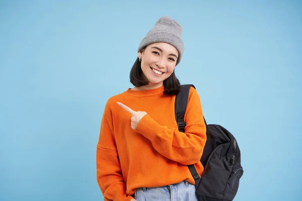 Enthusiastic asian woman, points finger left at banner, shows promo offer, smiles and looks happy, stands in hat with backpack, blue background.