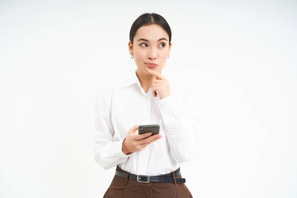 stock image Asian business woman, young manager holds smartphone and thinks, looks thoughtful aside, makes decision, stands over white background.