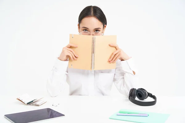 Korean woman hides her face behind notebook, peeks at camera, sits at desk with headphones and tablet, white background.