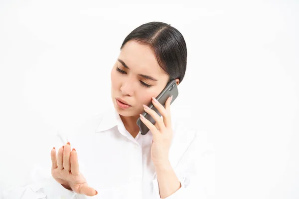 Young corporate employee, woman talks on mobile phone with lack on interest, looks at her nails while listens to person on telephone, white background.