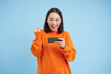 Cep telefonuyla video oyunu kazanan mutlu Koreli kız, elinde cep telefonu ve kutlama, uygulamada hedefe ulaşmak, mavi arka planda durmak.