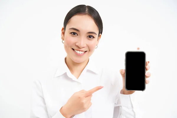 stock image Portrait of businesswoman showing smartphone screen, pointing at mobile phone and smiling, recommending application, standing over white background.