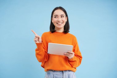 Tabletli modern Asyalı kadın, tanıtım teklifi, afiş için boş yere bakıyor, gülümsüyor ve reklamlara bakıyor, mavi arka plan.