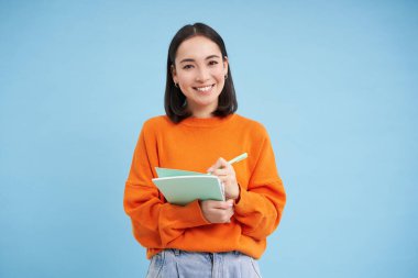 Güzel Asyalı kız, öğrenci not alıyor, yazıyor, derslere çalışıyor, öğreniyor, mavi arka planda duruyor..