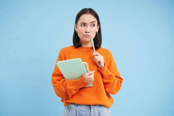 Asian girl, student with thinking face, pondering smth with serious face, holding pencil and notebooks, standing over blue background.
