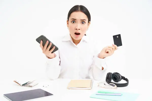 stock image Portrait of asian woman sits at table, holds credit card and smartphone, looks upset, disappointed by prices, white background.