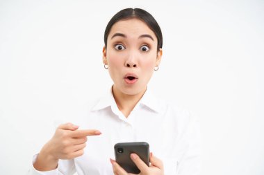 Mutlu satış elemanlarının yakın portresi, akıllı telefonlu başarılı kadın, telefonuna işaret ediyor ve şaşkın bir şekilde gülümsüyor, beyaz arka planda duruyor..