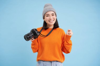 Dijital kameralı şık Asyalı kız fotoğraf çekiyor. Kadın fotoğrafçı gülümsüyor, mavi arka planda duruyor..