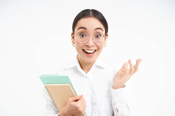 Portrait of corporate woman, asian woman in glasses, holds notebook and looks surprised, amazed by smth, stands over white background.