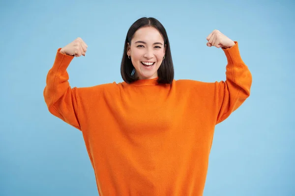 Strong and healthy people. Young asian woman shows her muscles, flexing biceps and looks confident, smiling pleased, in great shape, blue background.