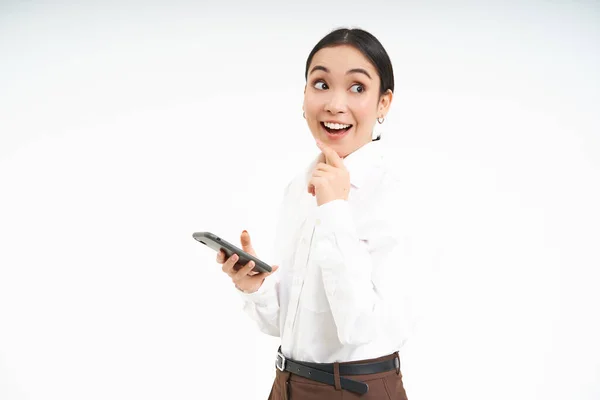 stock image Enthusiastic corporate woman holds smartphone, looks excited and thoughtful, uses social media, stands over white background.