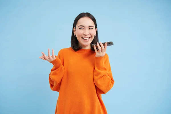 Happy asian woman records voice message, repeats words in language learning mobile app, talks in smartphone speakerphone, blue background.