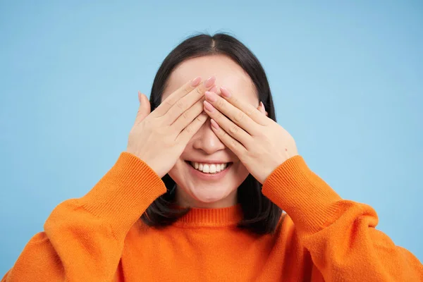 Close up of asian woman shuts her eyes and smiles blindfolded, waits for surprise, stands over blue background.