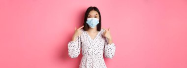 Coronavirus, sosyal uzaklık ve yaşam tarzı konsepti. Yüz maskesini işaret eden sevimli Asyalı kadın, kovid 19 'a karşı önlem almak istiyor, pembe arka planda duruyor..